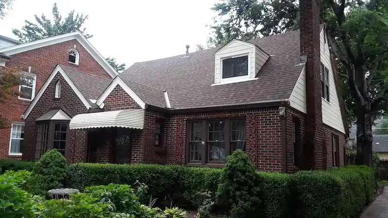A finished brick Tudor-style home with a striped awning over the front window and clipped hedges along the walk