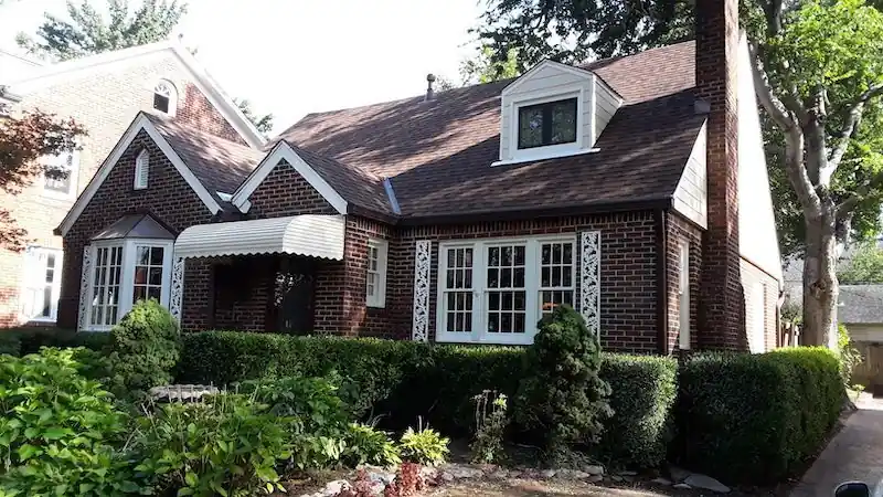 A finished brick Tudor-style home with a cream-sided dormer, striped awnings and clipped hedges